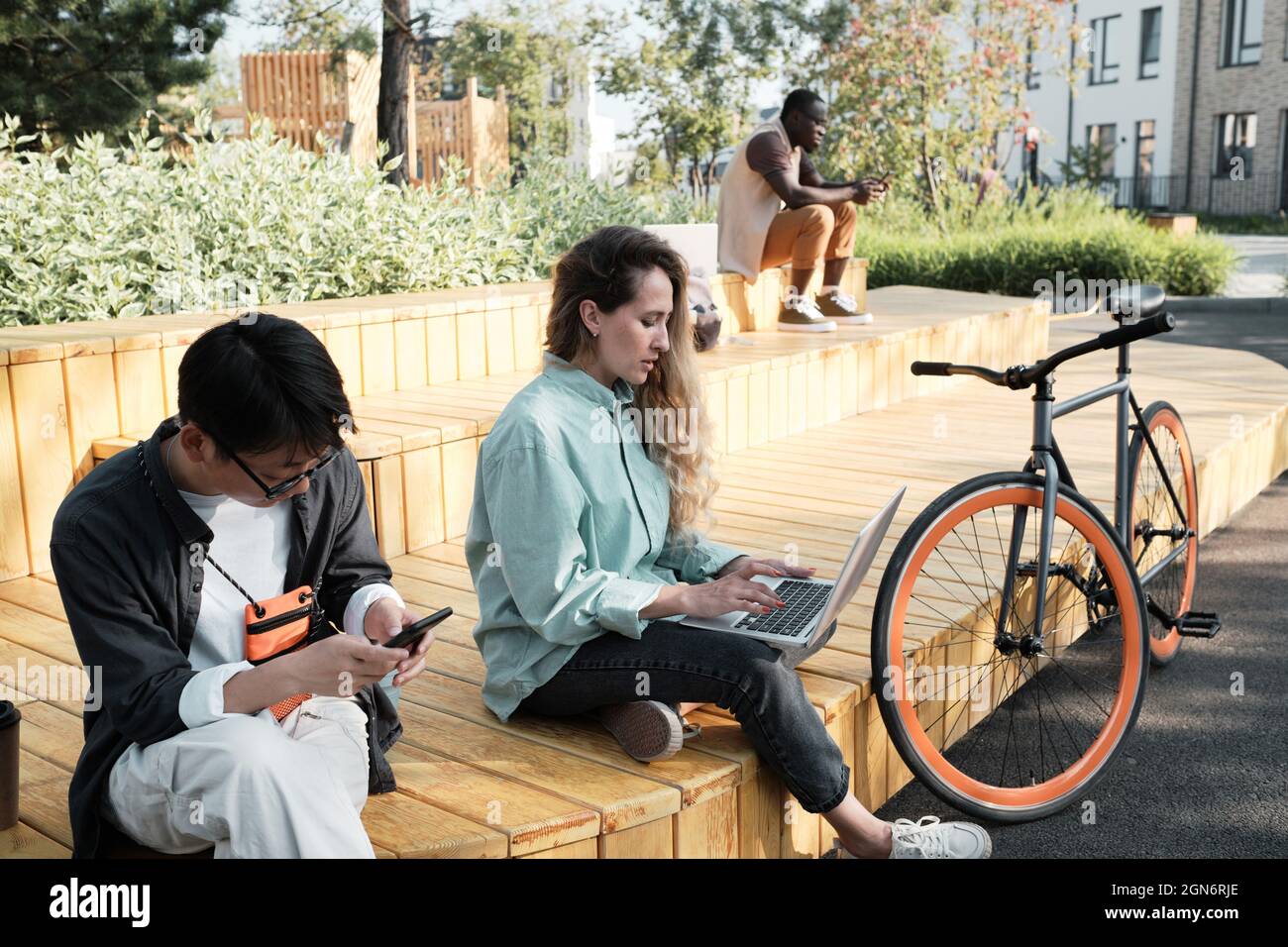Groupe de trois jeunes gens multiethniques passent la journée d'été dehors assis dans un parc urbain moderne travaillant à distance ou surfant sur Internet sur leurs gadgets Banque D'Images