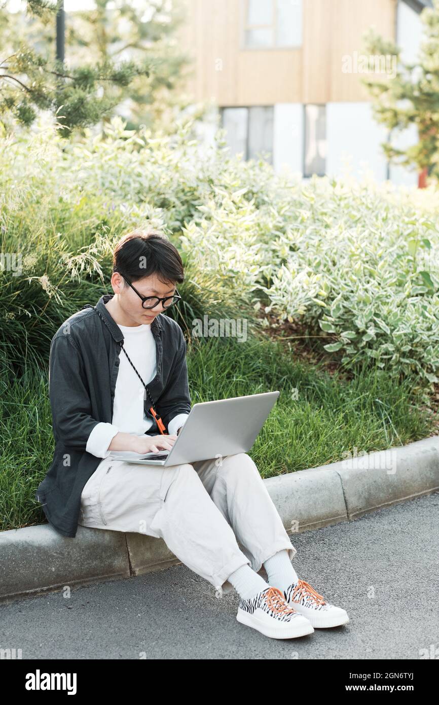 Prise de vue verticale d'une jeune femme asiatique sérieuse portant des vêtements décontractés assis sur un trottoir à l'extérieur, axée sur un travail indépendant à l'aide d'un ordinateur portable Banque D'Images