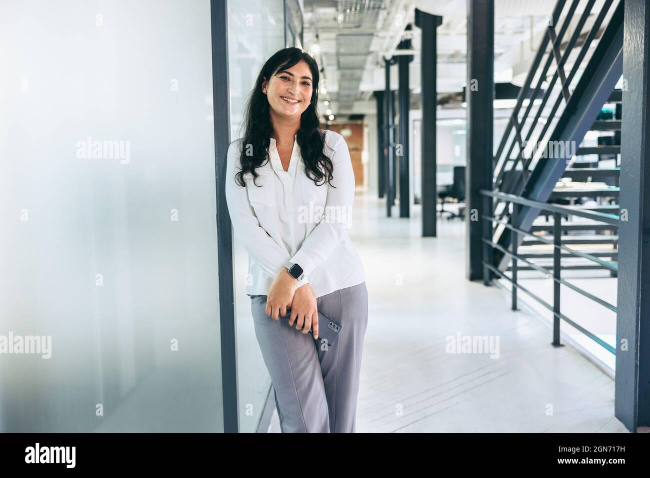 Bonne femme d'affaires de taille moyenne souriante à l'appareil photo. Jeune femme d'affaires tenant un smartphone tout en restant seule dans un bureau moderne. Busi réussi Banque D'Images Bonne femme d'affaires de taille moyenne souriante à l'appareil photo. Jeune femme d'affaires tenant un smartphone tout en restant seule dans un bureau moderne. Busi réussi Banque D'Images