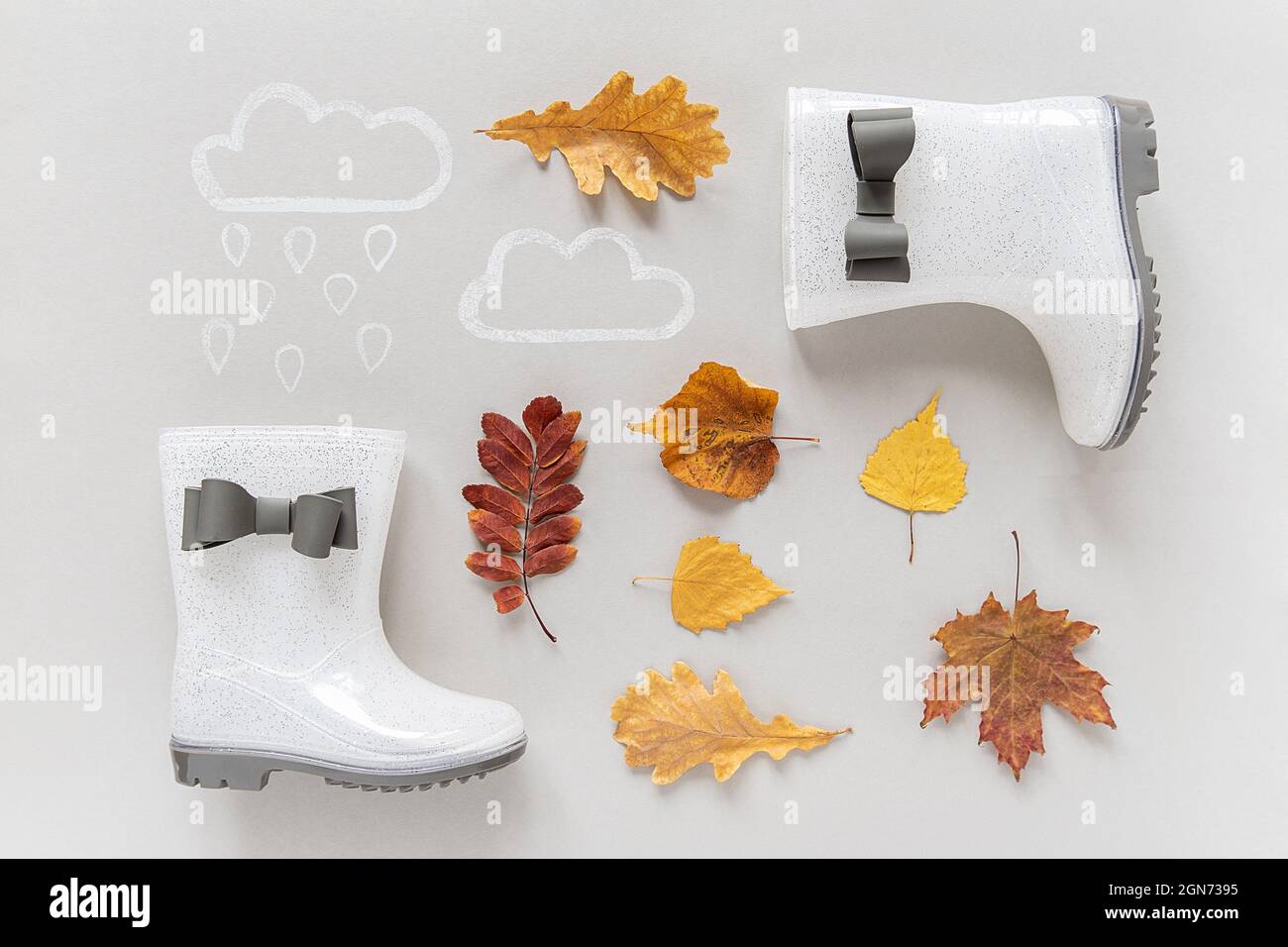 Concept d'automne créatif. Bottes de pluie en caoutchouc blanc brillant avec arcs, branche d'arbre avec feuilles d'automne et nuages de pluie peints sur fond gris. Banque D'Images