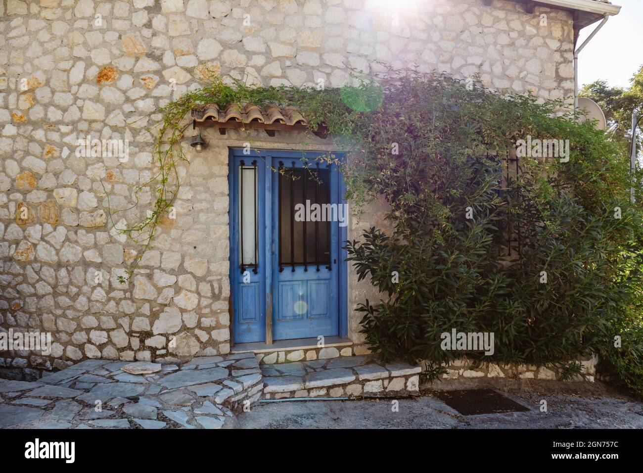 Le soleil brille au-dessus du toit traditionnel grec de la maison avec des murs en pierre, des volets de portes bleues et des branches vertes. Voyage d'été dans la Méditerranée traditionnelle vi Banque D'Images
