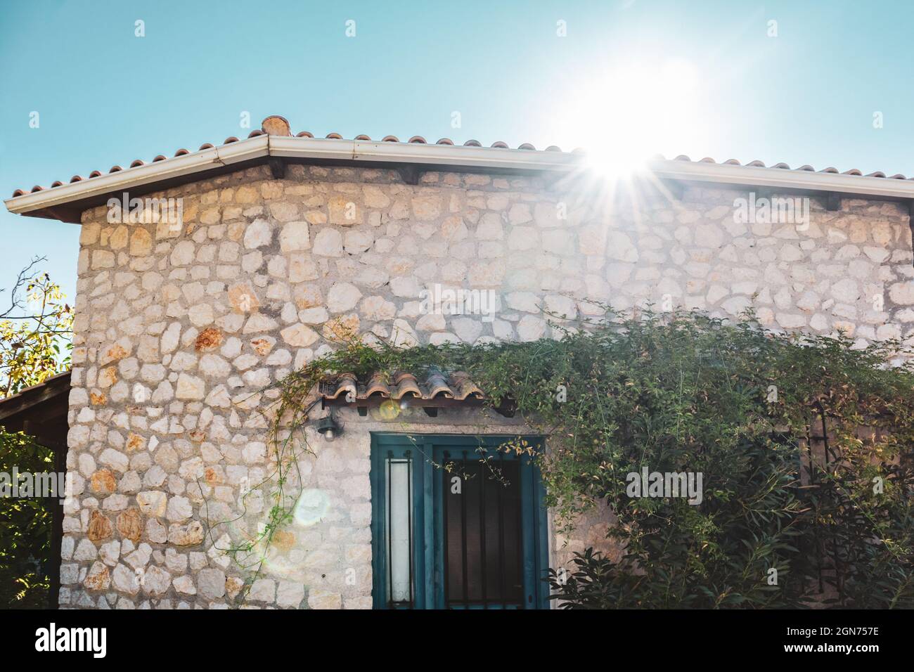 Le soleil brille au-dessus du toit traditionnel grec de la maison avec des murs en pierre, des volets de portes bleues et des branches d'arbres vertes. Voyage d'été en Méditerranée traditionnelle Banque D'Images