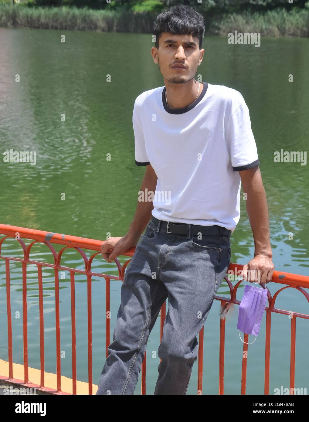 Un beau jeune homme nord-indien portant un t-shirt blanc et un Jean noir et tenant le masque de visage à portée de main avec assis sur la barrière de sécurité au bord du lac et regardant la caméra Banque D'Images