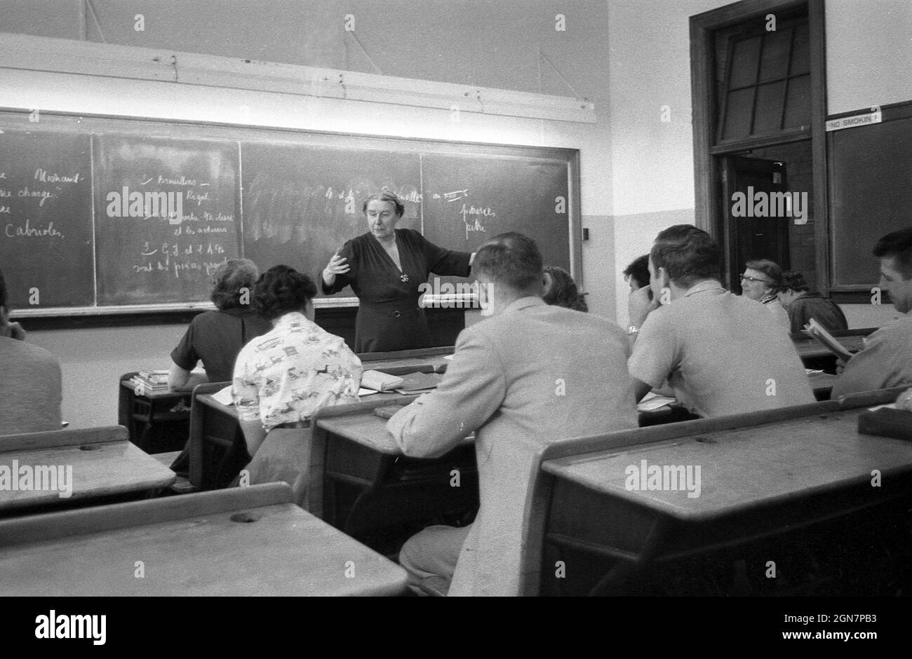 Années 1970, historique, une femme enseignante debout à un tableau noir enseignant une classe française à un groupe d'étudiants matures assis sur des bancs en bois dans une salle de classe d'université, Allemagne. Banque D'Images