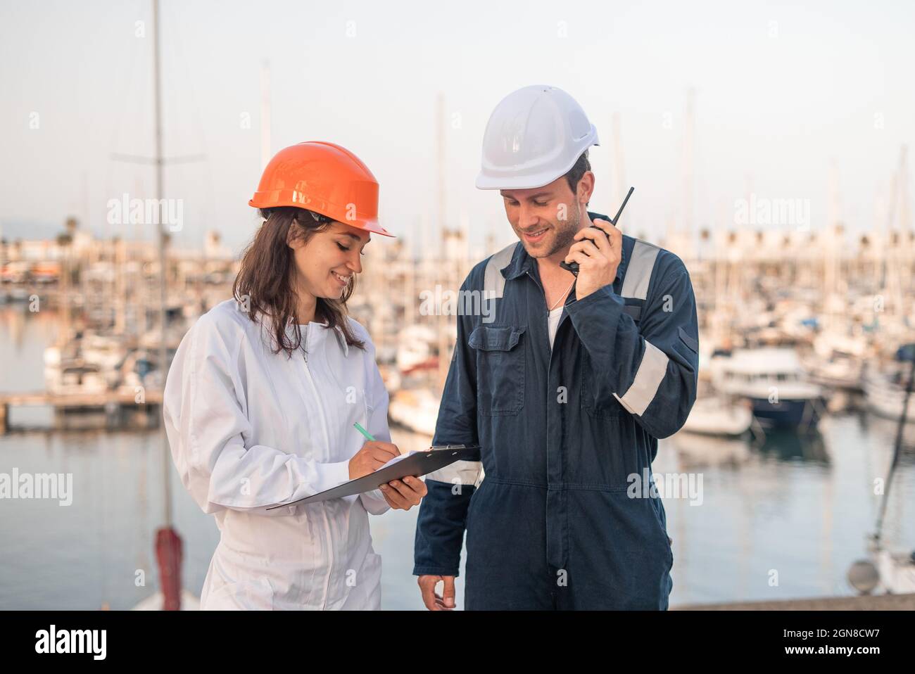 Une jeune femme inspecteur positive prend des notes sur le presse-papiers lors de la préparation du rapport lors d'une réunion avec un ingénieur mâle avec un talkie-walkie dans le port maritime Banque D'Images