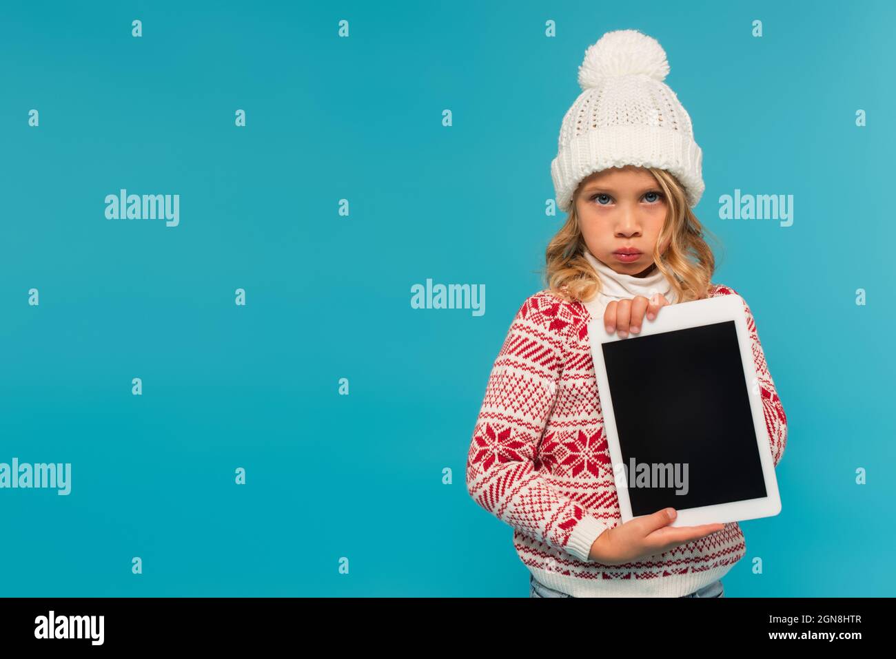 enfant ennuyé dans un chapeau chaud et un chandail tenant une tablette numérique avec écran vierge isolé sur bleu Banque D'Images