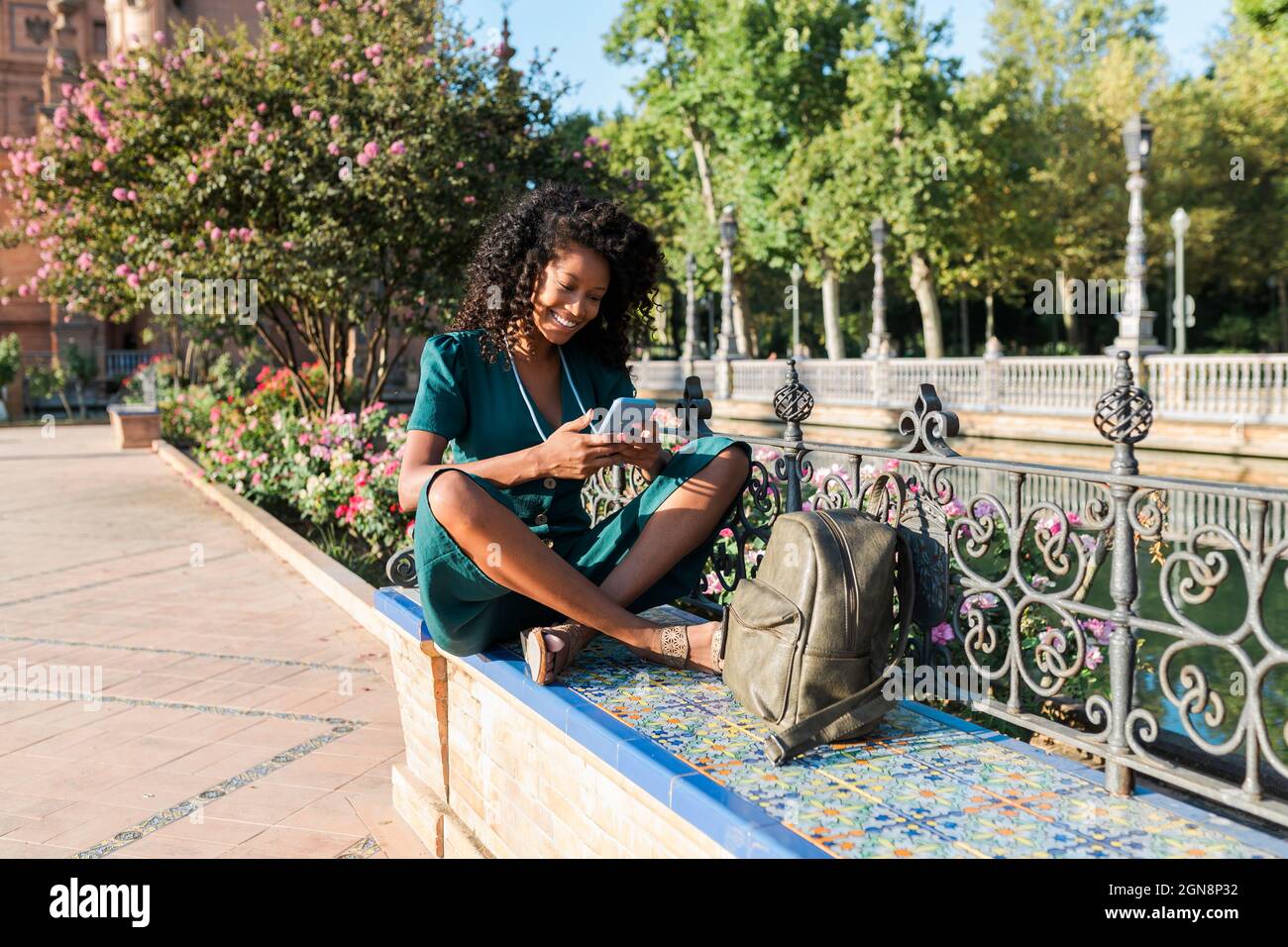 Souriante touriste féminine utilisant le téléphone portable tout en étant assise à pieds croisés sur le banc de la Plaza de Espana, Séville, Espagne Banque D'Images