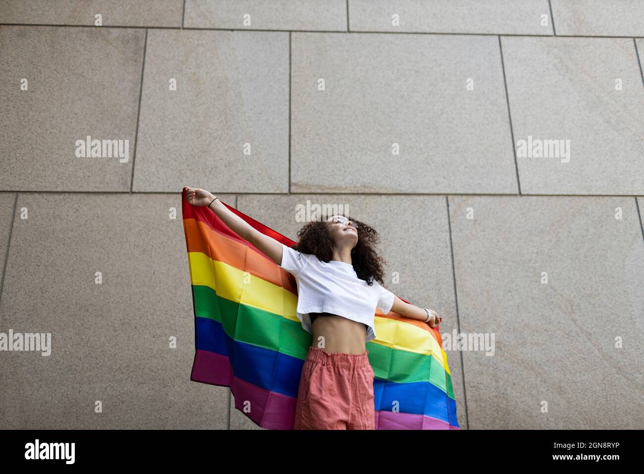 Femme tenant le drapeau arc-en-ciel en se tenant devant le mur Banque D'Images