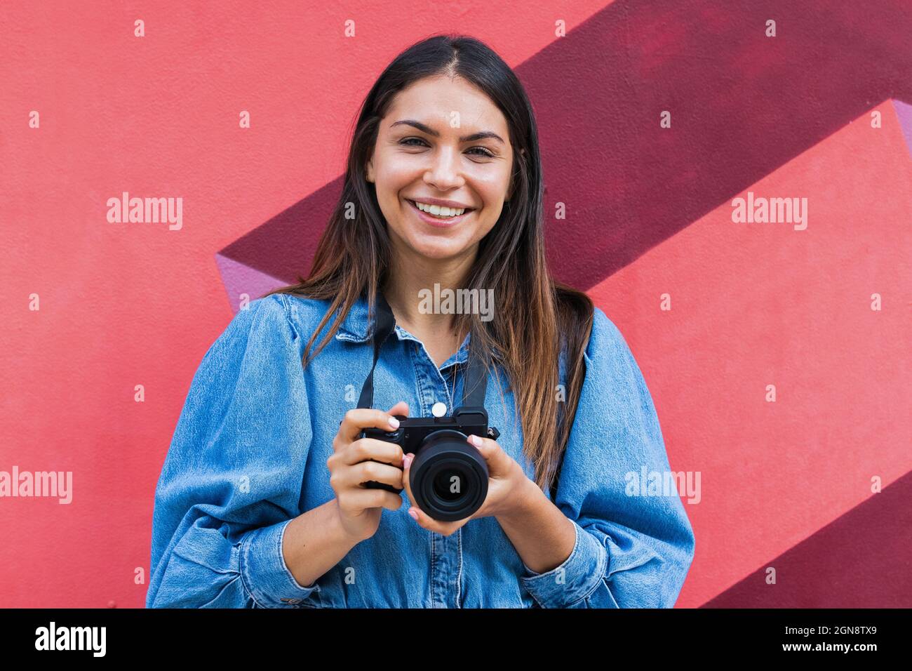 Femme souriante travailleur indépendant avec caméra debout devant le mur Banque D'Images