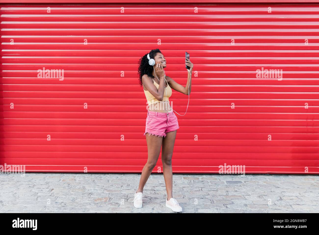Une jeune femme souriante prend son selfie à travers un smartphone tout en écoutant de la musique devant l'obturateur rouge Banque D'Images Une jeune femme souriante prend son selfie à travers un smartphone tout en écoutant de la musique devant l'obturateur rouge Banque D'Images