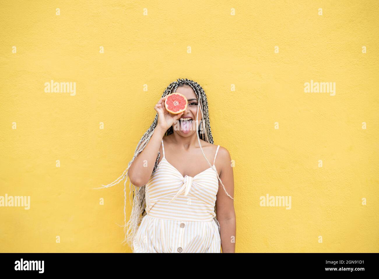 Femme qui colle la langue tout en tenant les agrumes devant les yeux par un mur jaune Banque D'Images