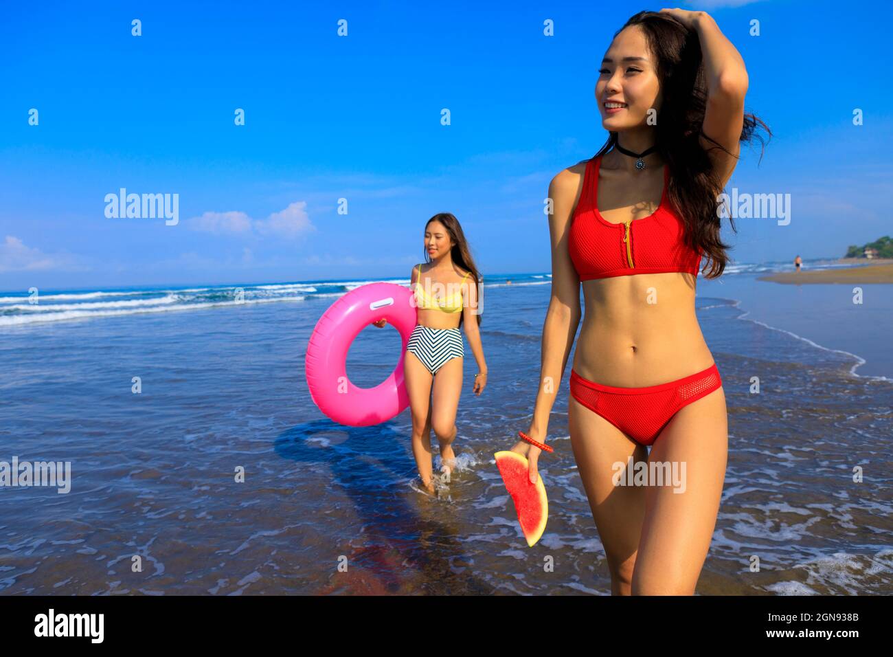 Femme souriante tenant la pastèque tandis qu'une femme avec la nage flottant marchant dans l'eau le jour ensoleillé Banque D'Images