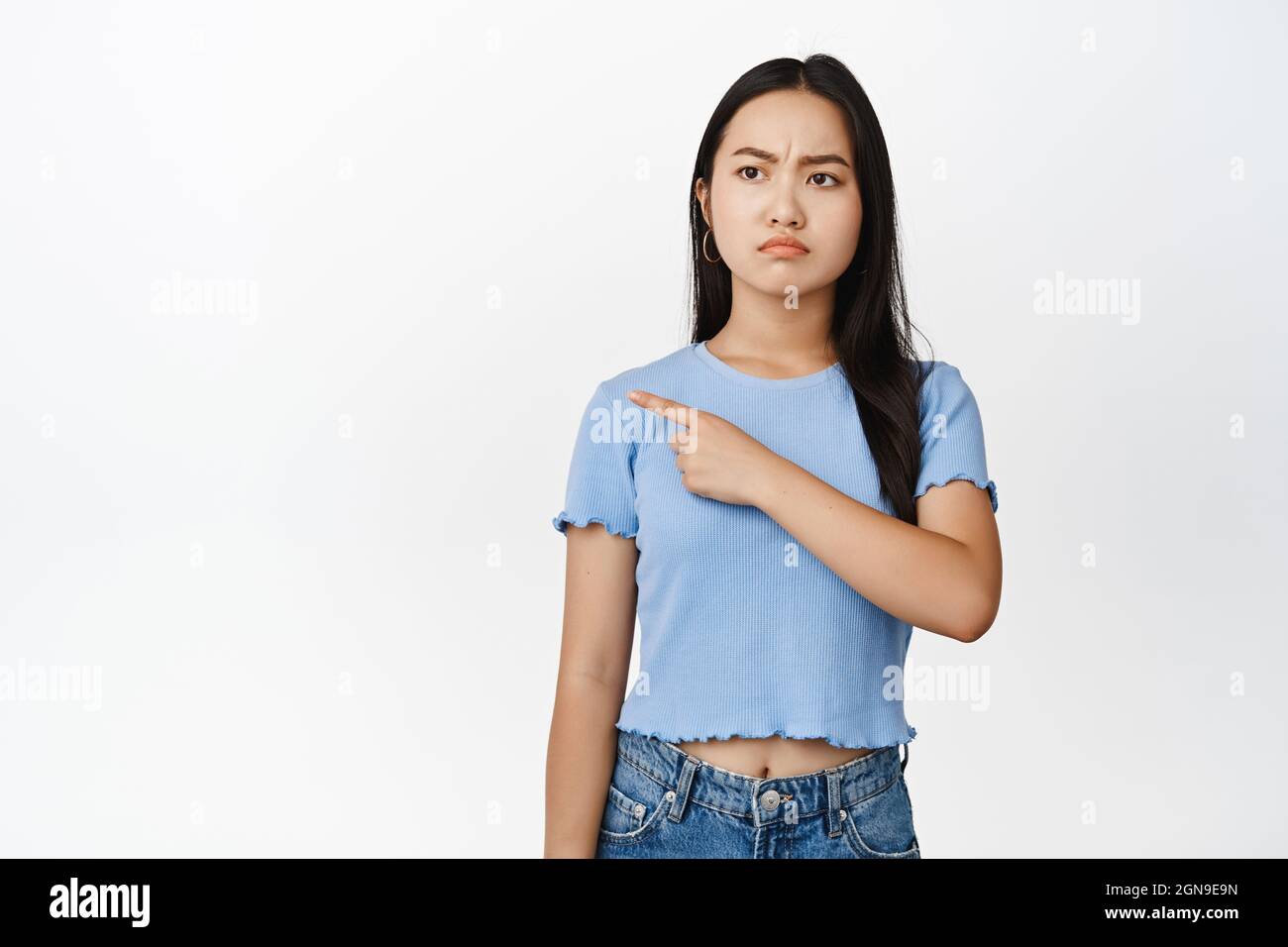 Une jeune fille asiatique en colère pointant vers la gauche avec un visage frogné et grognant, se lasser et se tenir debout sur un fond blanc Banque D'Images