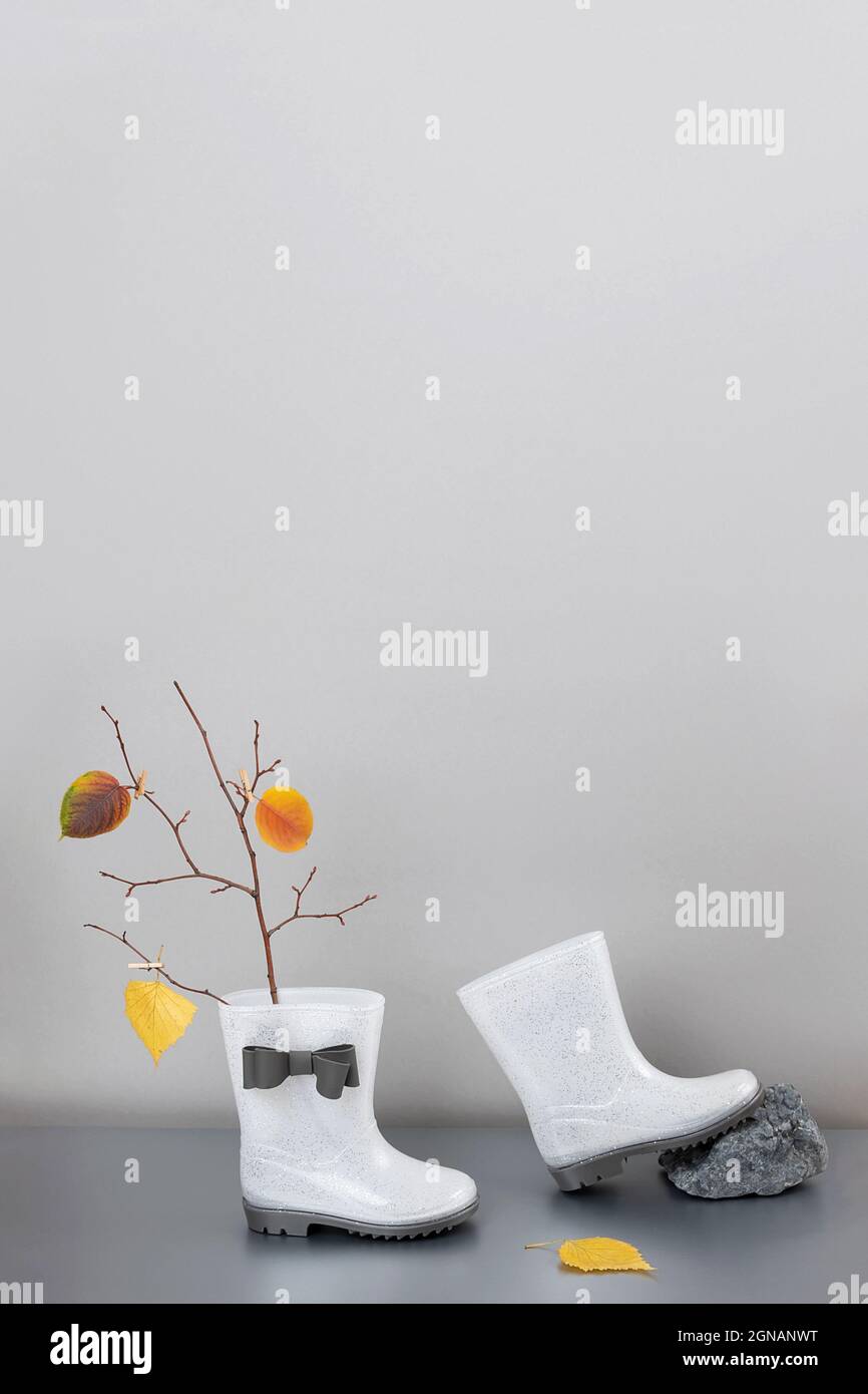 Concept d'automne créatif. Bottes de pluie en caoutchouc blanc brillant avec arcs, branches d'arbre avec feuilles d'automne sur fond gris. Copier l'espace. Banque D'Images