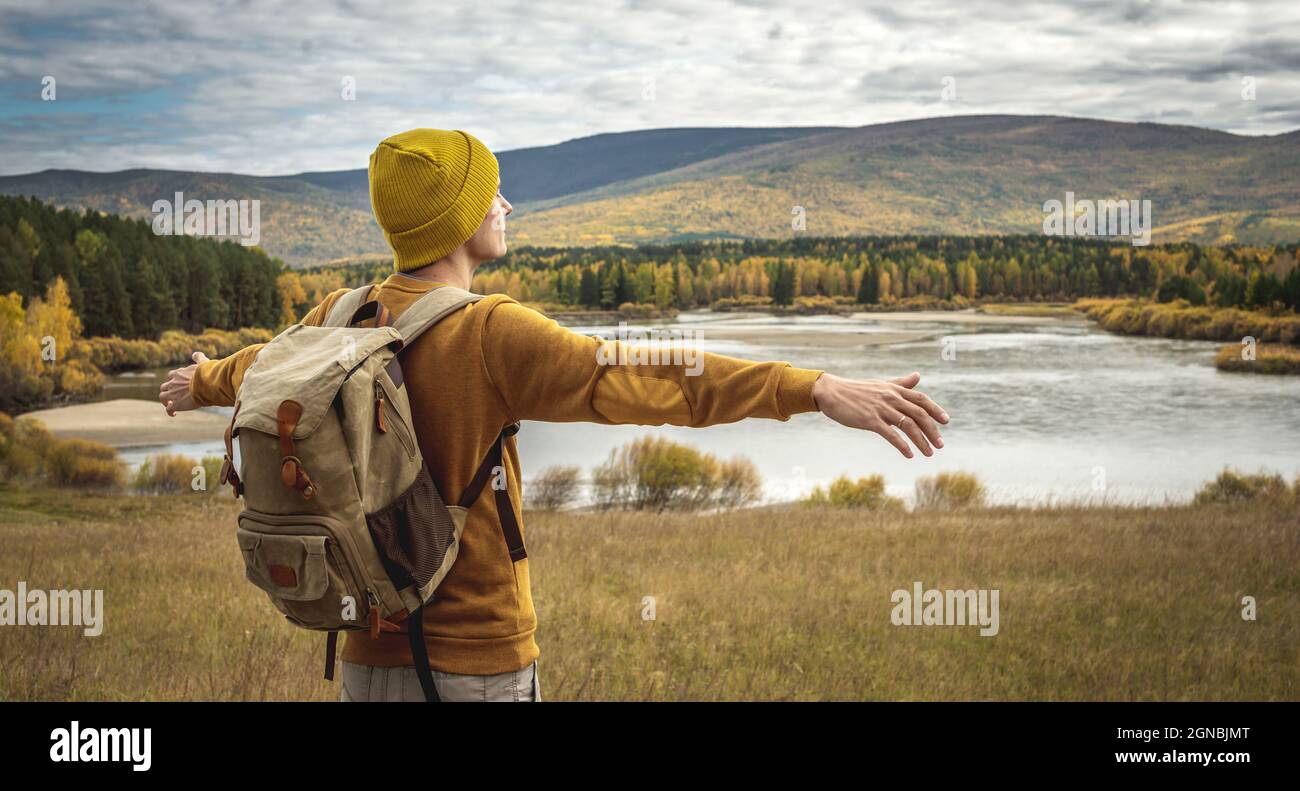 Touriste dans un chapeau jaune et chandail avec un sac à dos est debout avec ses bras étendus sur les côtés en face d'une rivière, la forêt dorée d'automne Banque D'Images