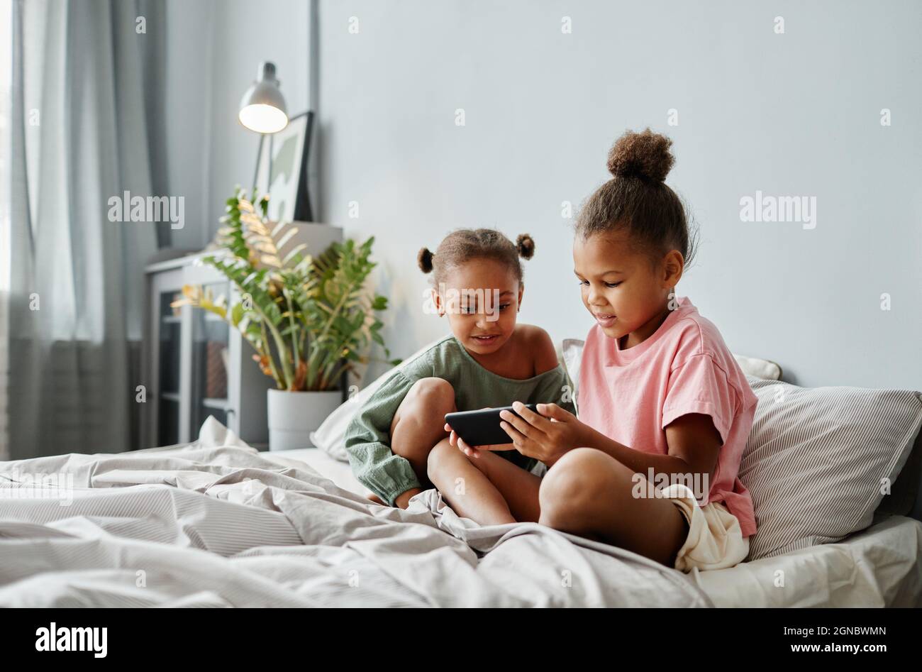 Portrait de deux filles afro-américaines utilisant un smartphone ensemble sur un lit dans une pièce confortable, espace de copie Banque D'Images