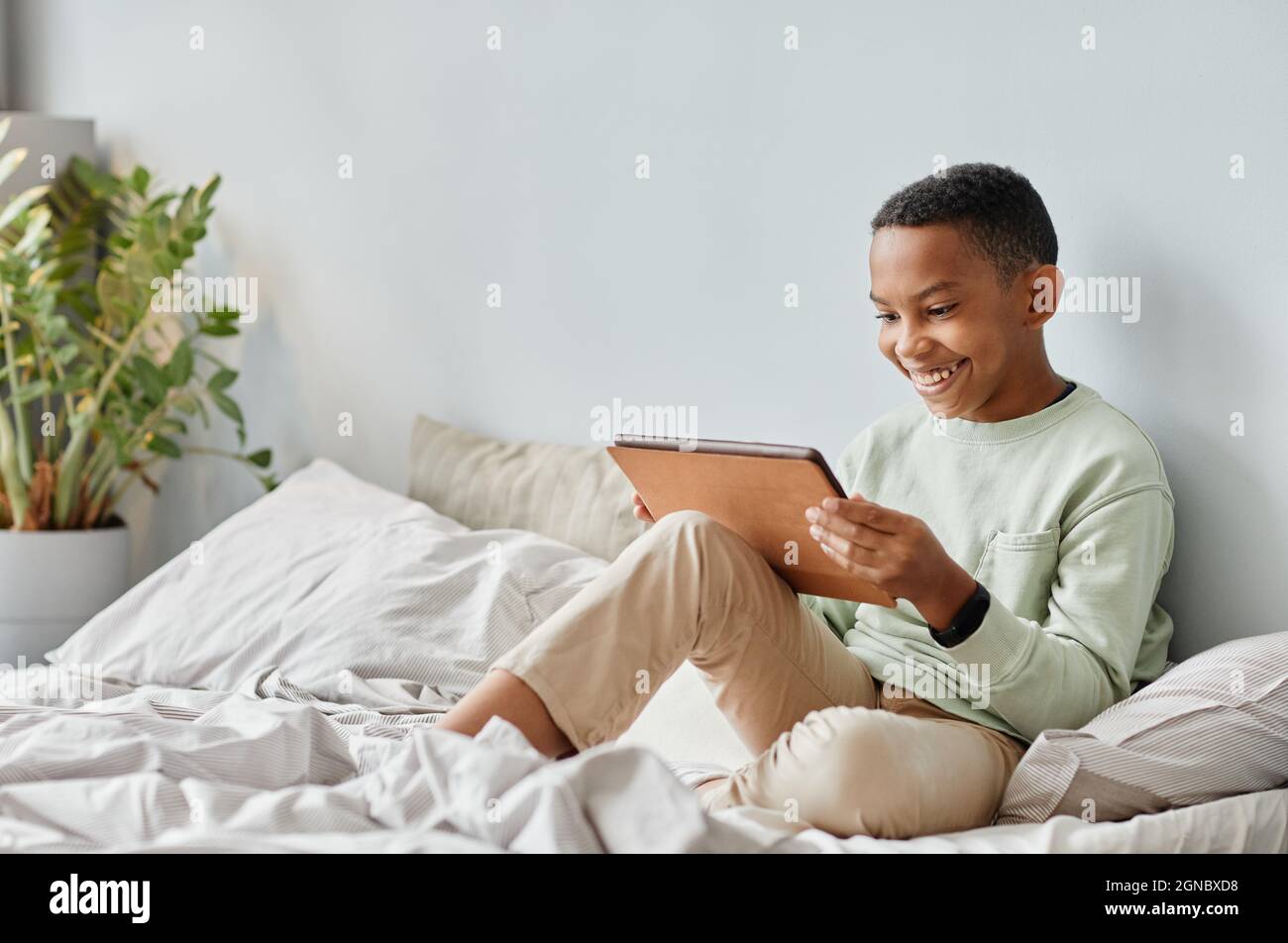 Portrait complet d'un adolescent afro-américain souriant utilisant une tablette tout en étant assis sur un lit dans une pièce confortable, espace de copie Banque D'Images