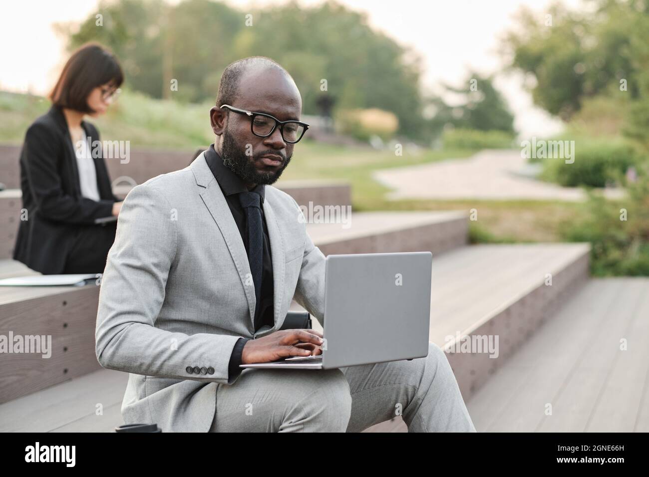 Portrait horizontal moyen d'un homme afro-américain sérieux et mature avec une barbe sur le visage portant un élégant costume assis dans un parc urbain à l'aide d'un ordinateur portable Banque D'Images