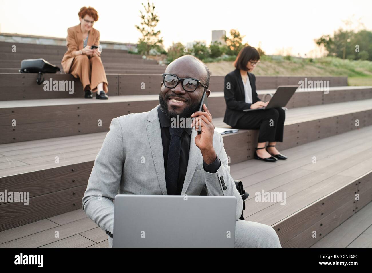 Portrait horizontal moyen d'un costume d'homme d'affaires afro-américain à barbe en toute confiance assis sur un banc à l'extérieur avec un ordinateur portable parlant au téléphone avec un co-worke Banque D'Images