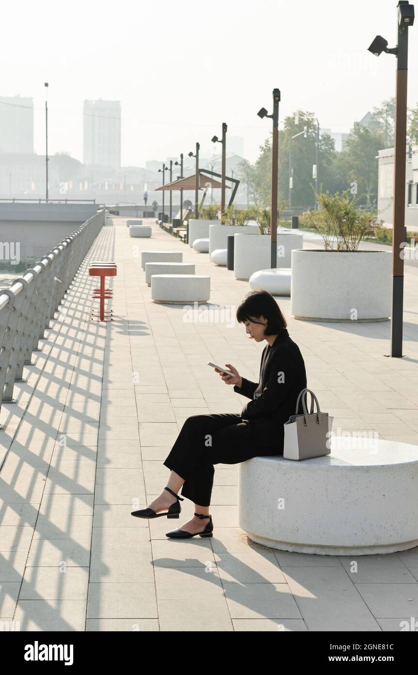 Plan vertical long de la belle jeune femme asiatique portant une tenue élégante et élégante assis à l'extérieur surfer sur Internet sur un smartphone, espace de copie Banque D'Images