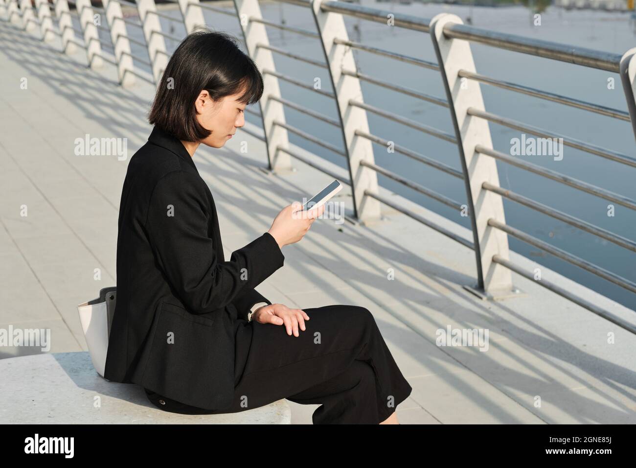 Prise de vue horizontale d'une jeune femme asiatique sérieuse portant des vêtements élégants et tendance, assise à l'extérieur, en vérifiant son plan de travail sur son smartphone, son espace de copie Banque D'Images