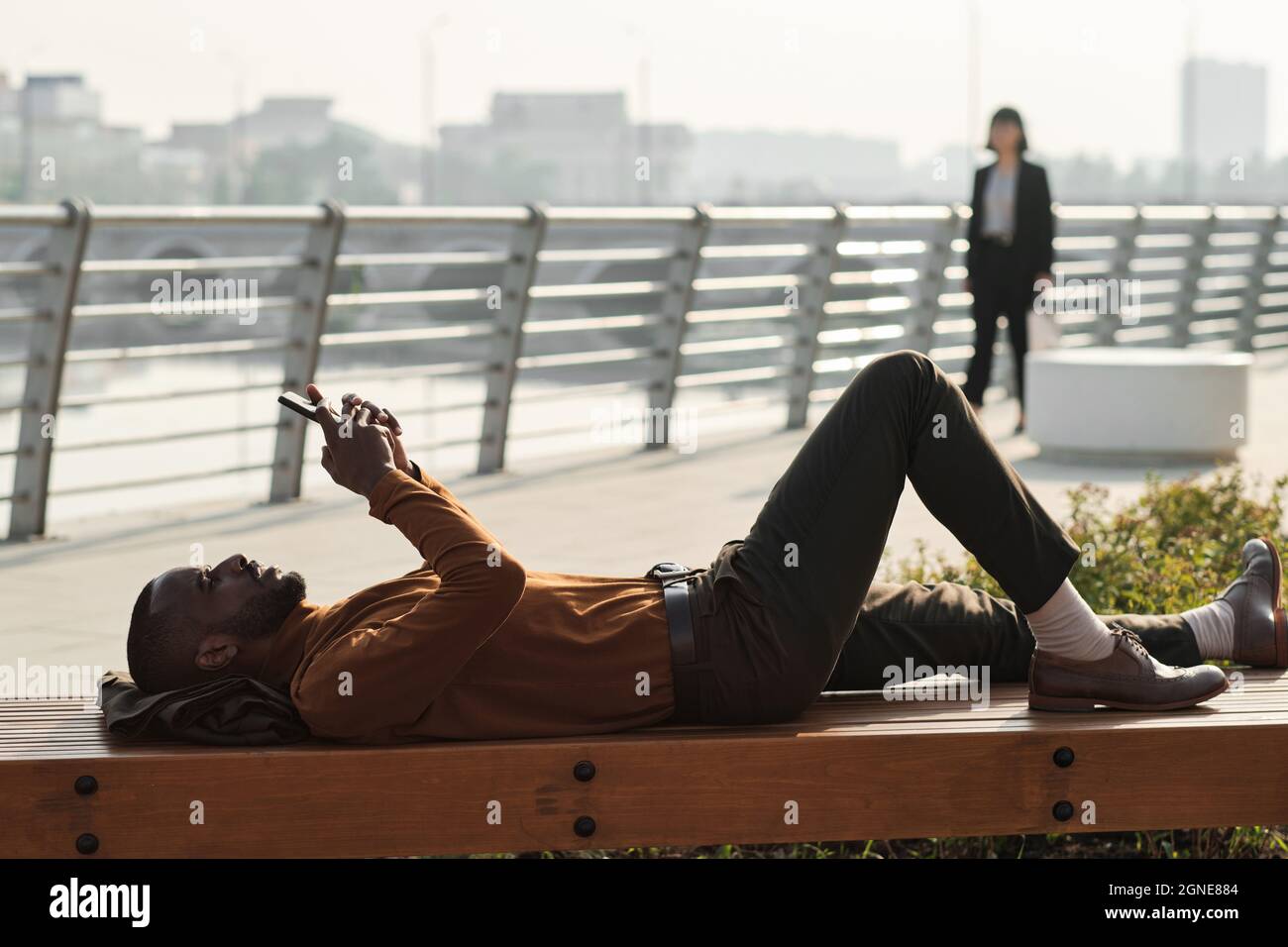 Vue latérale horizontale prise de vue d'un homme afro-américain moderne se reposant sur un banc en bois à l'extérieur en regardant quelque chose sur un smartphone Banque D'Images