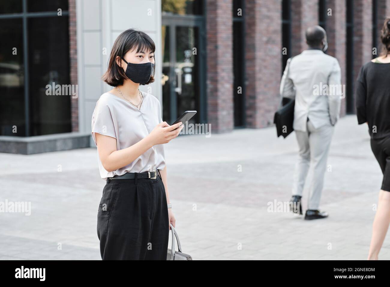 Portrait horizontal moyen d'une jeune femme asiatique portant un masque de protection sur le visage tenant un smartphone debout dans le quartier des affaires en attendant son co-wo Banque D'Images