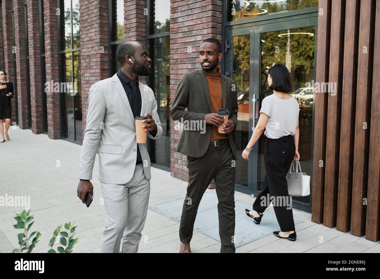 Plan horizontal moyen long de deux beaux hommes noirs portant des tenues tendance tenant des tasses avec le café marchant et parlant pendant la pause Banque D'Images