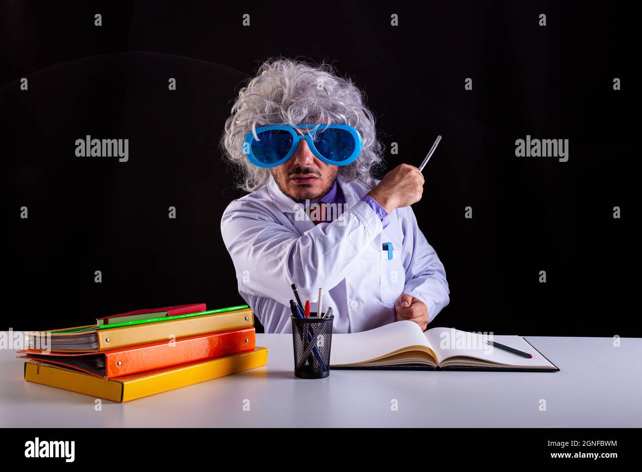Professeur de science fou en manteau blanc avec des cheveux unkempt dans des lunettes drôles d'oeil assis au bureau tenant une baguette pour pointer au tableau noir Banque D'Images