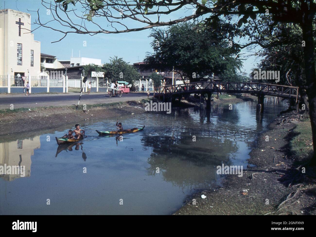 De jeunes enfants thaïlandais pagayent des canoës traditionnels le long d'un canal à Bangkok, en Thaïlande, en passant par une église chrétienne. 1968. Une passerelle en bois traverse le canal . Banque D'Images