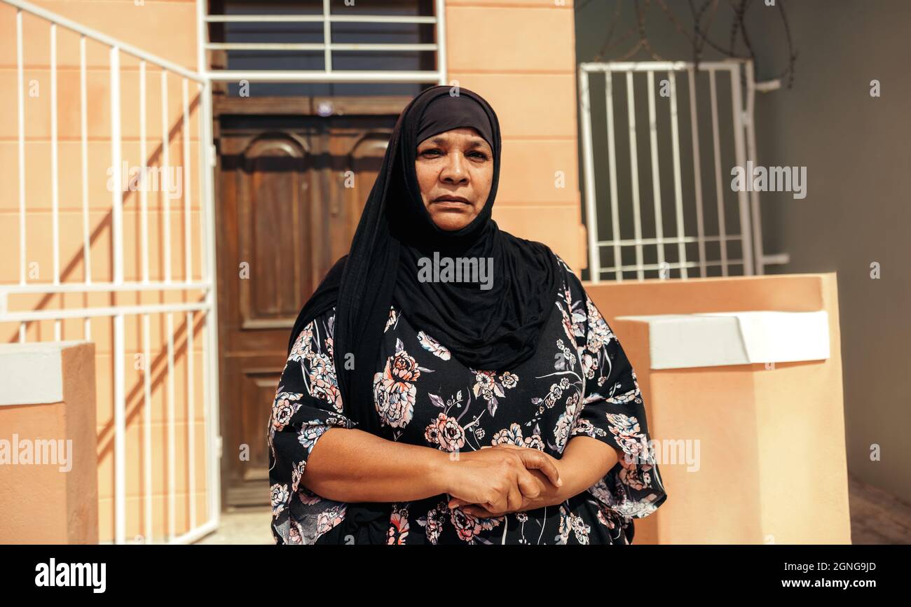 Femme musulmane confiante regardant la caméra tout en se tenant devant sa maison pendant la journée. Femme d'âge moyen confiante portant un hijab et Banque D'Images Femme musulmane confiante regardant la caméra tout en se tenant devant sa maison pendant la journée. Femme d'âge moyen confiante portant un hijab et Banque D'Images