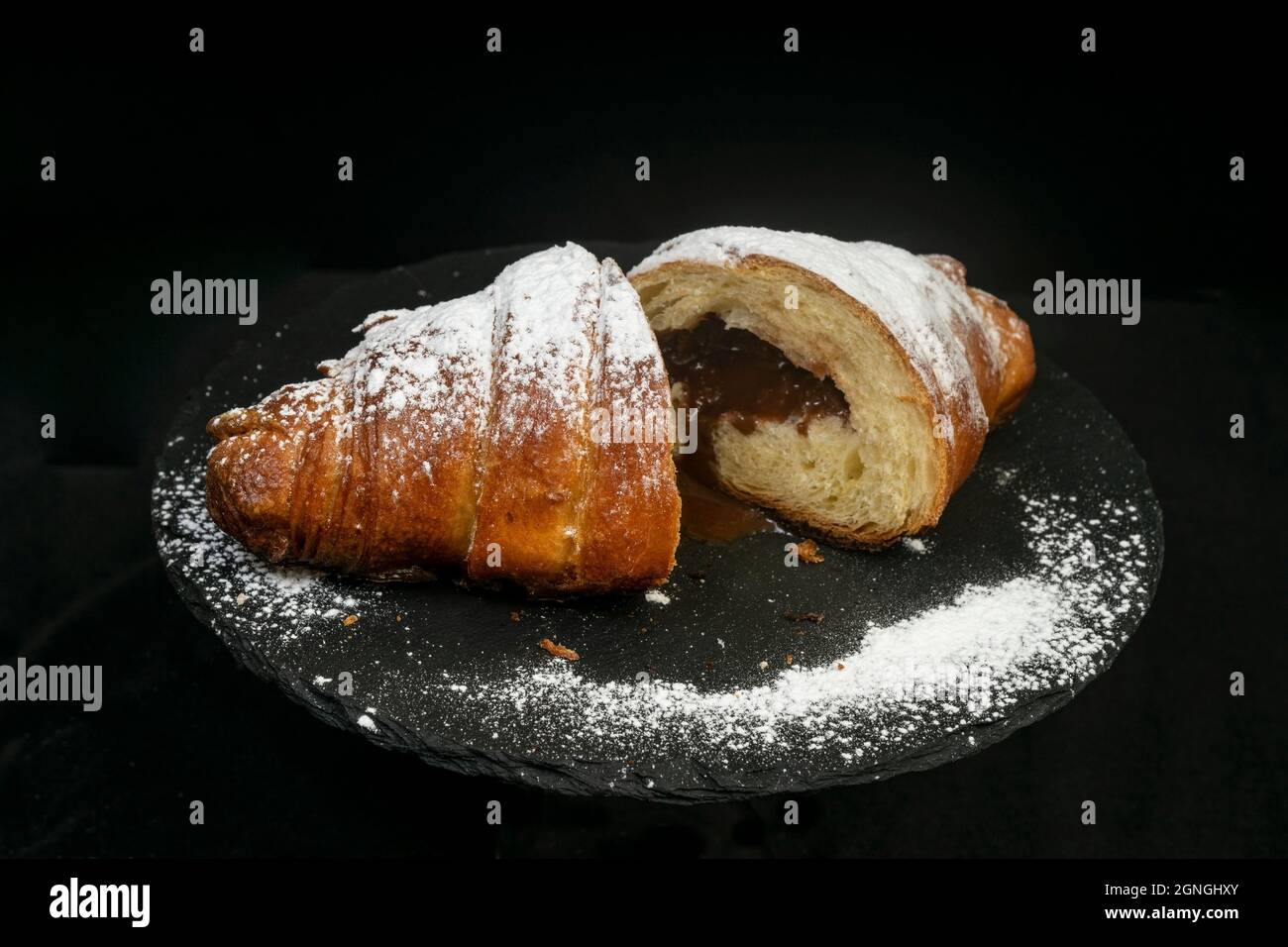 2 moitiés d'un croissant au chocolat, poudrées de sucre, sur une tuile de pierre noire, isolée sur fond noir. Banque D'Images