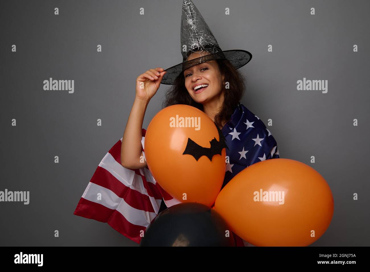 Joyeuse femme de course mixte dans un chapeau de sorcier, enveloppée d'un drapeau américain, contient des boules d'air orange colorées, sourit un sourire éclatant en regardant l'appareil photo. Halloween c Banque D'Images