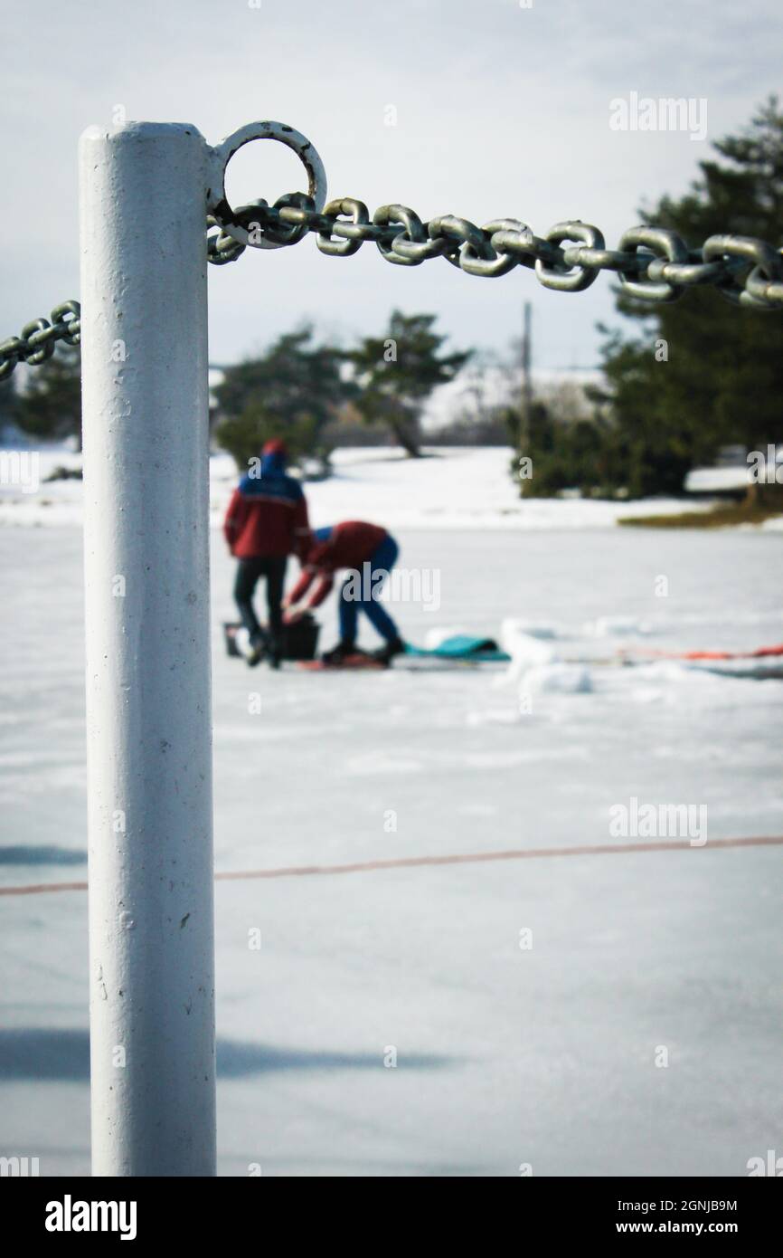 Deux hommes à distance qui font un trou dans la glace pour faire du verglas | deux personnes qui coupent le trou dans le lac gelé avec un poteau de pont au premier plan Banque D'Images