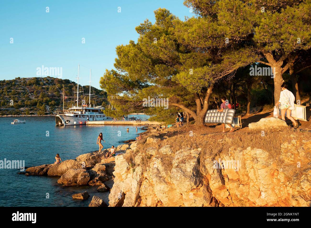 Podvrske, Murter, Croatie - 26 août 2021 : pins au bord de la plage rocheuse, personnes transportant des accessoires de plage et bateau amarré au coucher du soleil d'été Banque D'Images