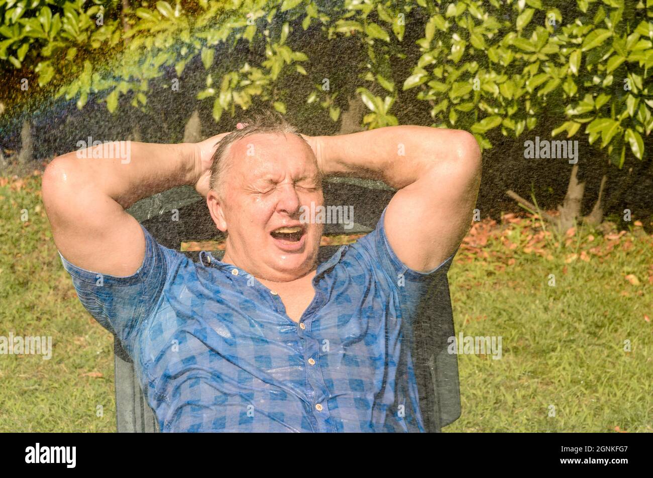 Portrait d'un homme mûr heureux sous l'eau des éclaboussures lors d'une journée chaude dans le jardin. Banque D'Images