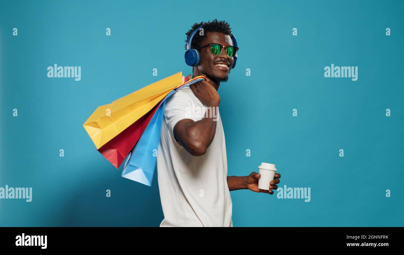 Homme heureux avec des sacs à main dansant après la frénésie de shopping. Personne joyeuse avec des lunettes de soleil écoutant de la musique sur les écouteurs tout en tenant des sacs avec des vêtements à vendre achat le vendredi noir Banque D'Images