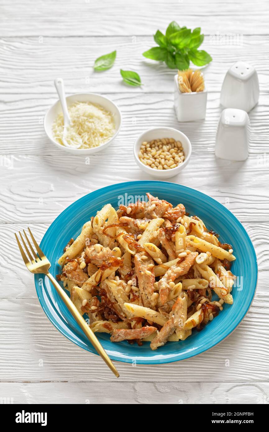 poulet alfredo pasta penne à l'oignon caramélisé arrosé de pignons de pin et de parmesan râpé sur une assiette bleue sur une table en bois blanc, à la verticale Banque D'Images