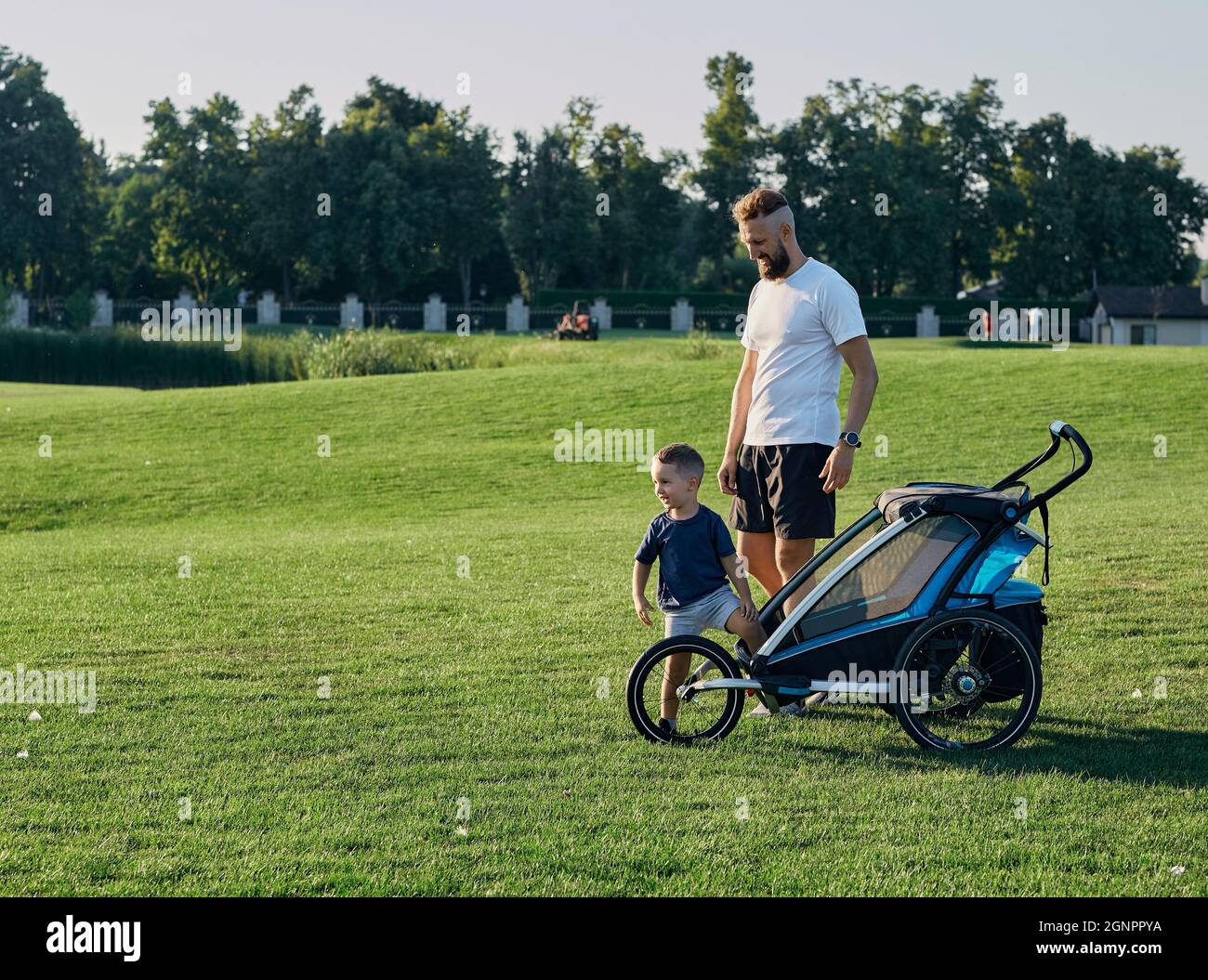Le père et son petit fils marchent dans le parc de la ville ensemble sur l'herbe verte avec la poussette de bébé. Paternité, papa et enfant. Fête des pères Banque D'Images