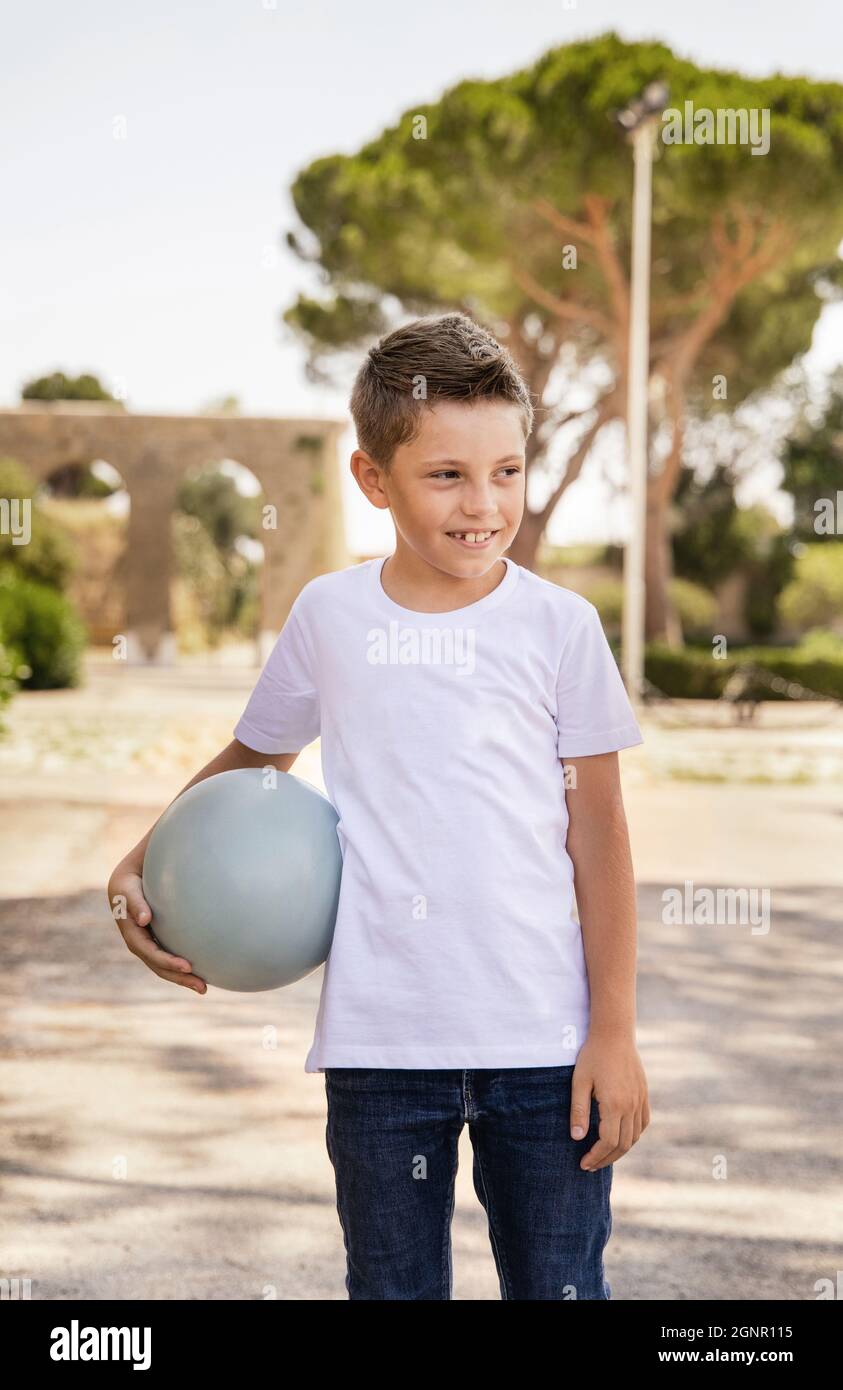 Joyeux garçon d'avant-garde dans un t-shirt blanc tenant le ballon à l'extérieur. Mignon enfant faisant du sport, appréciant jouer au football et s'amuser dans le parc. Passe-temps actifs, Banque D'Images