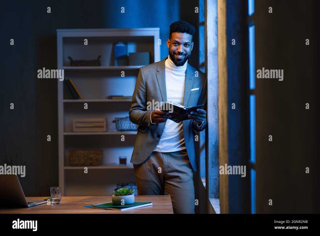 Jeune homme d'affaires afro-américain avec journal à l'intérieur dans le bureau la nuit, regardant la caméra. Banque D'Images