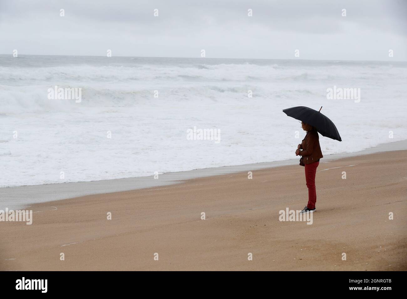 Femme avec parapluie noir debout devant le rivage de la mer un jour de tempête et de pluie. Nazaré. Portugal. Banque D'Images