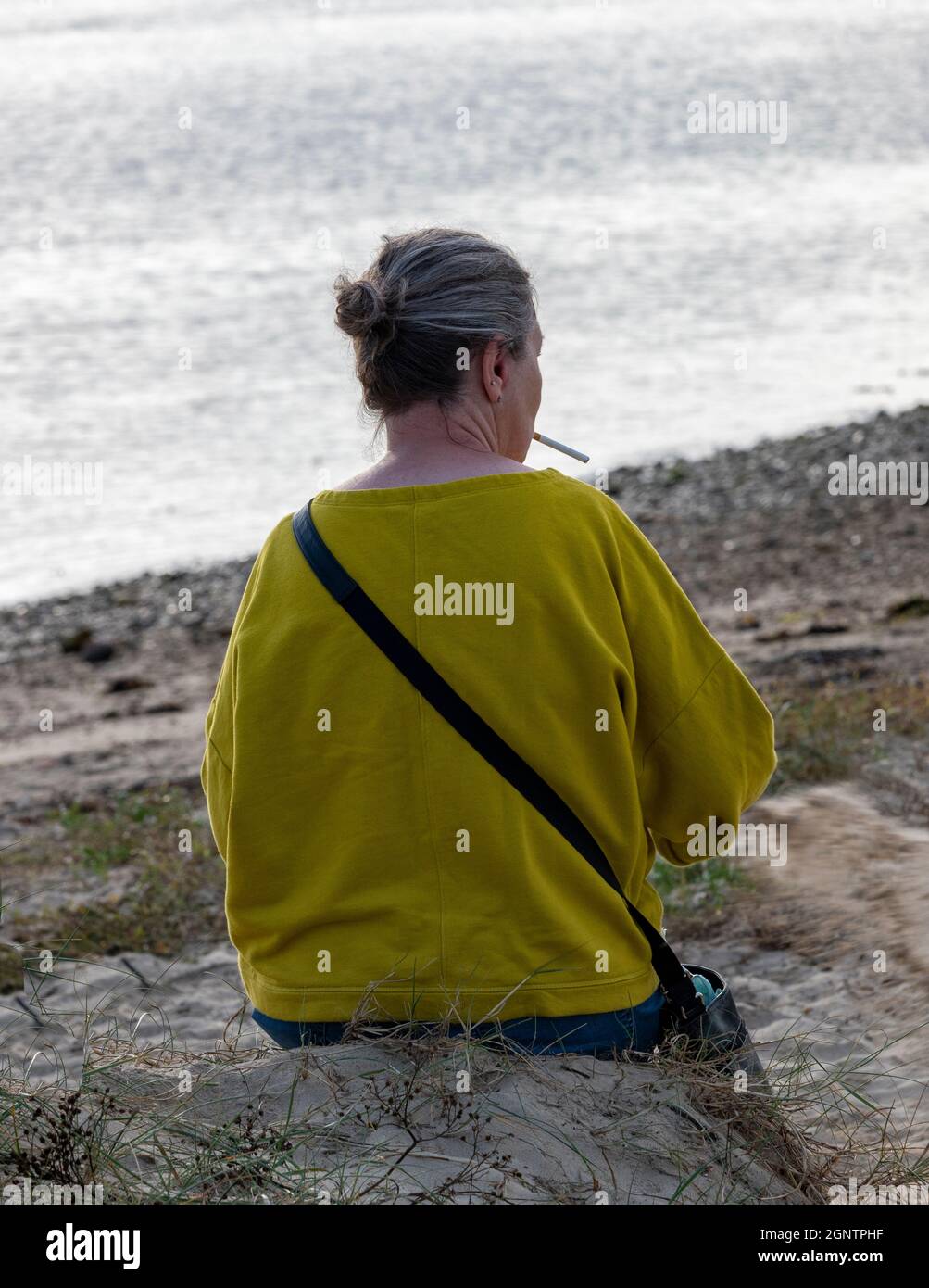 femme d'âge moyen assise sur une plage portant un cavalier jaune et fumant une cigarette. fumeur à la station balnéaire fumant une cigarette sur la plage. Banque D'Images