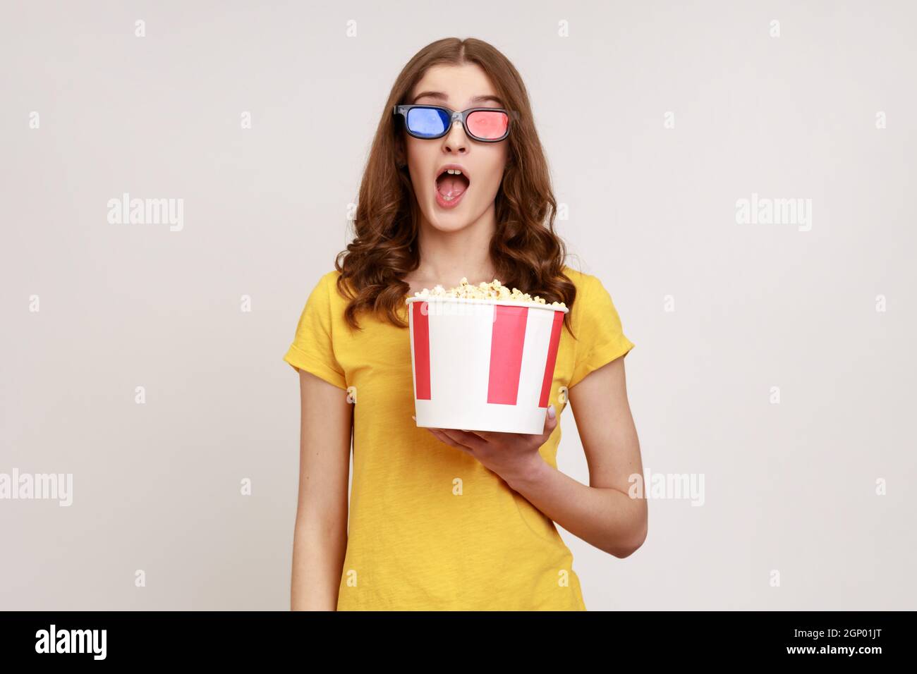 Une jeune femme stupéfiante en lunettes 3d imax regardant un film, tient du  pop-corn, regarde un film incroyable, garde la bouche ouverte, porte un  T-shirt jaune. Prise de vue en studio isolée
