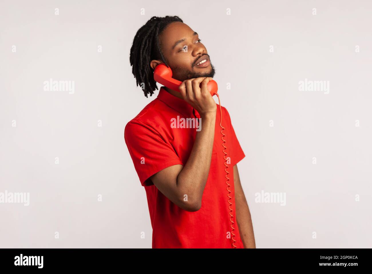 Vue latérale d'un homme avec des dreadlocks et une barbe portant un T-shirt rouge de style décontracté, passer des appels sur un téléphone fixe, regarder l'appareil photo avec un sourire crasseux. Prise de vue en studio isolée sur fond gris. Banque D'Images