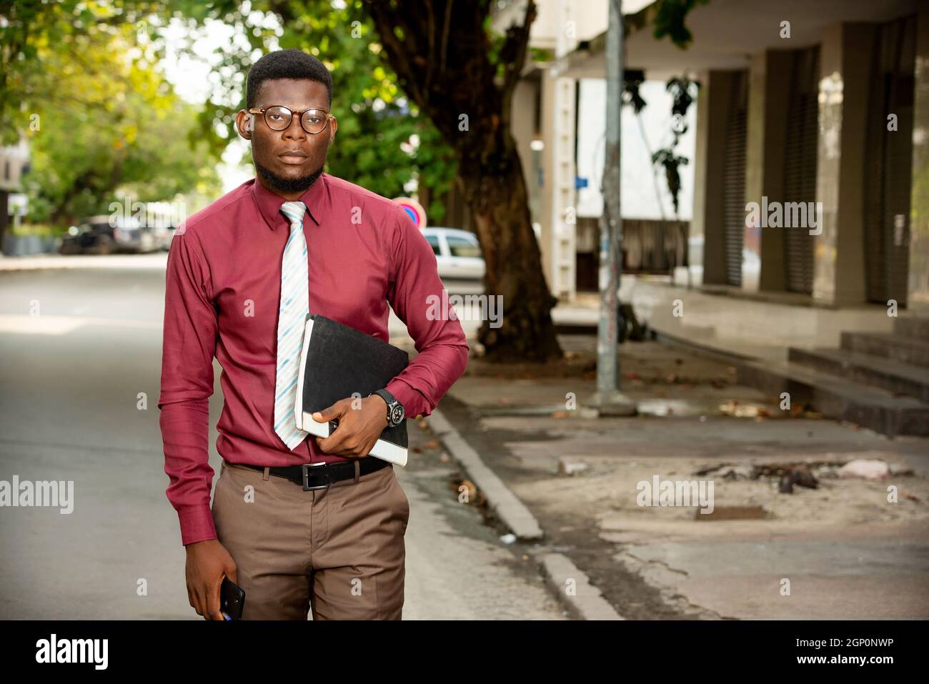 Beau homme africain qui se promette dans la rue et qui tient des documents.  Il porte une chemise rouge bordeaux très élégante avec cravate et lunettes  Photo Stock - Alamy
