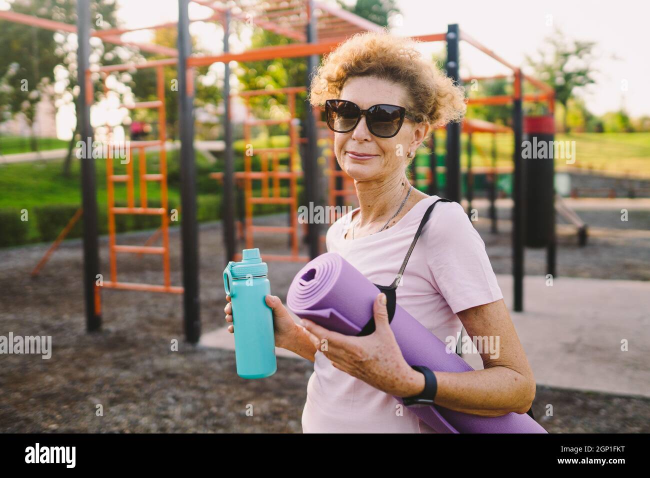 Femme active senior avec tapis de fitness et bouteille d'eau dans les mains marchant sur l'entraînement salle de gym extérieure. Bonne femme d'âge mûr se promenant dans un terrain de sport Banque D'Images