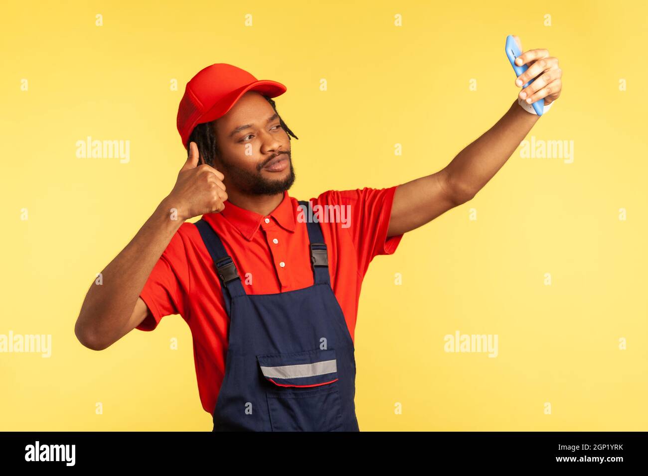 Beau travailleur avec la barbe portant l'uniforme ayant appel vidéo avec le client, montrant le pouce vers le haut, parlant du travail parfait, exécuter l'ordre. Studio d'intérieur isolé sur fond jaune. Banque D'Images