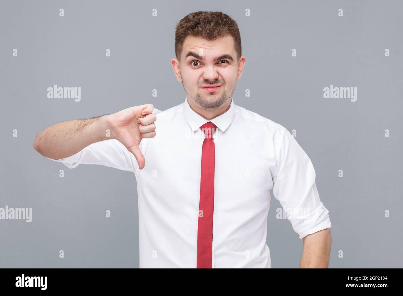 Portrait d'un jeune homme insatisfait en chemise blanche et cravate debout, pouces vers le bas et regardant l'appareil photo avec déplaît. Intérieur isolé sur fond gris. Banque D'Images