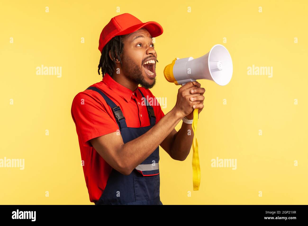 Portrait d'un homme à barbe portant une combinaison bleue et une casquette rouge criant d'annoncer des réductions dans l'industrie des services ou de protester. Studio d'intérieur isolé sur fond jaune. Banque D'Images